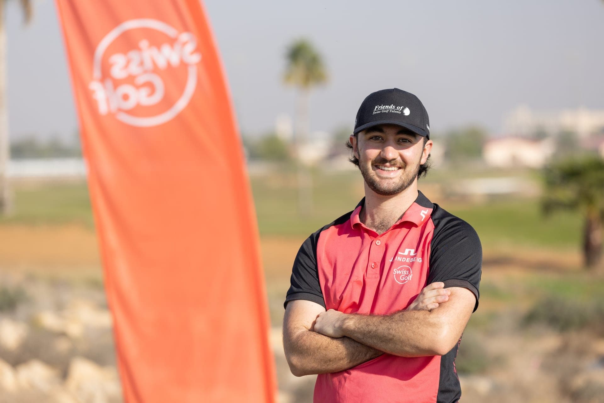 Lachender Golfprofi Mauro Gilardi mit verschränkten Armen neben einer Swiss-Golf-Fahne